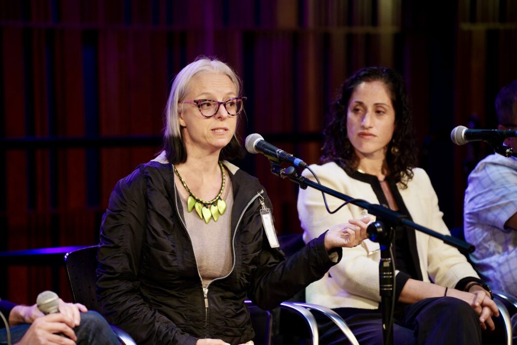 A woman speaks into a microphone. She appears to be on a panel, sitting next to another woman.