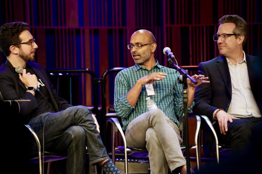 A male panelist in the center of the frame speaks to a man on his left, while another man listens on his right.