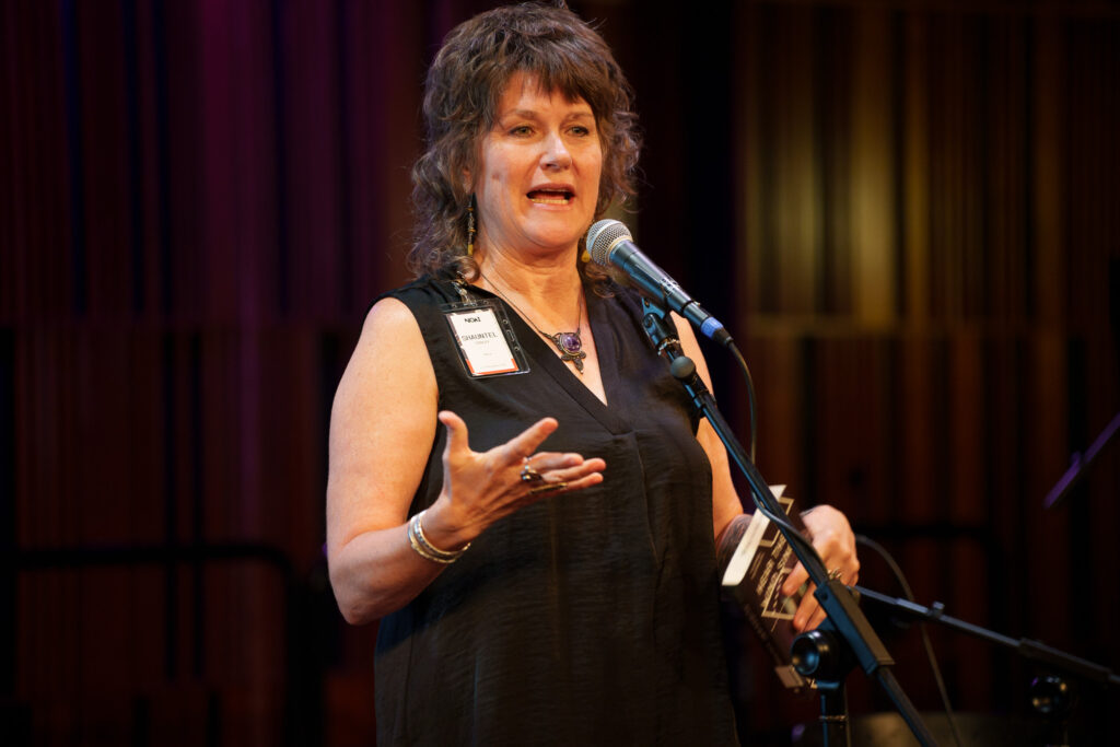 A female speeaker is speaking into a microphone. She appears to be mid-sentence.