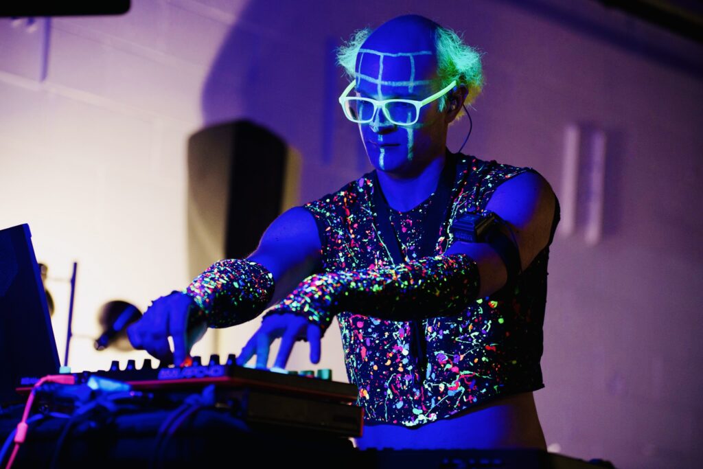 An electronic musician, painted in neon ink, turns knobs on his instrument.