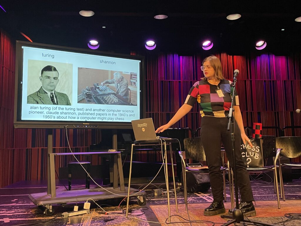 A speaker stands with her hand to a laptop. Behind her is a screen with a slide with two photographs, slightly out of focus.