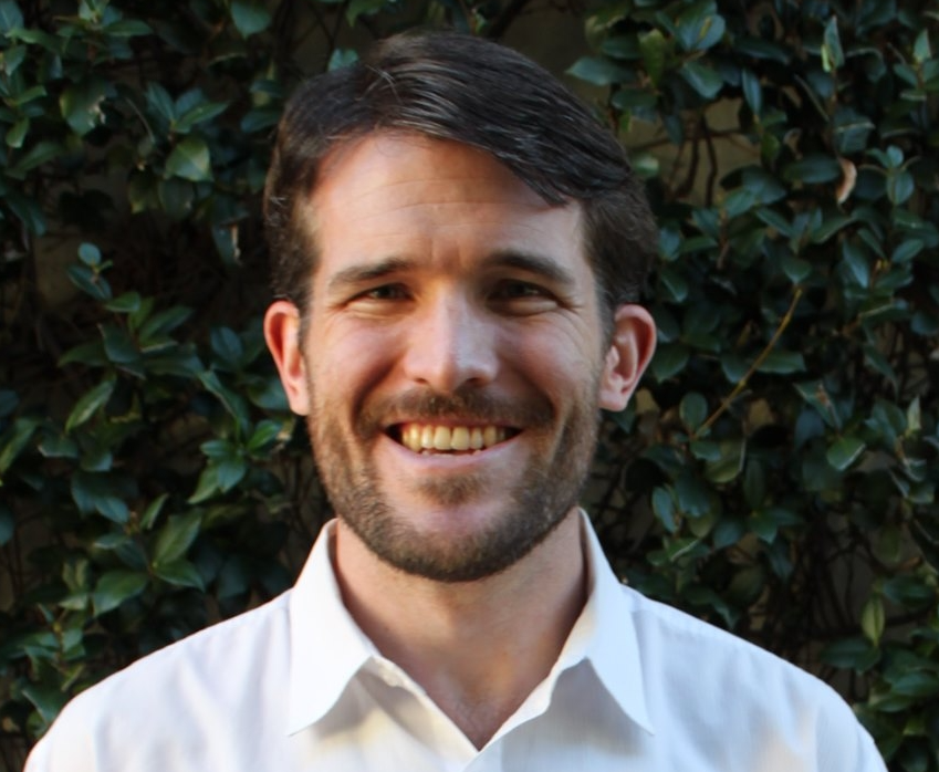 head shot of man a smiling man in a dress shirt against an ivy clad wall in the background